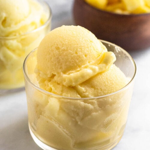 A glass cup with scoops of pineapple dole whip in it. Behind it is another glass cup filled and a bowl of frozen pineapple.