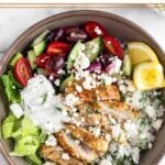 Greek chicken bowl with chopped romaine, tomato cucumber salad, herby rice, sliced chicken thighs, tzatziki, and feta and garnished with dill and 2 lemon wedges.