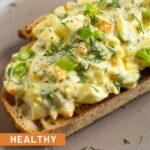 A close up of healthy egg salad on a piece of toast on a brown plate. It is topped with sliced green onions, dill, and black pepper.