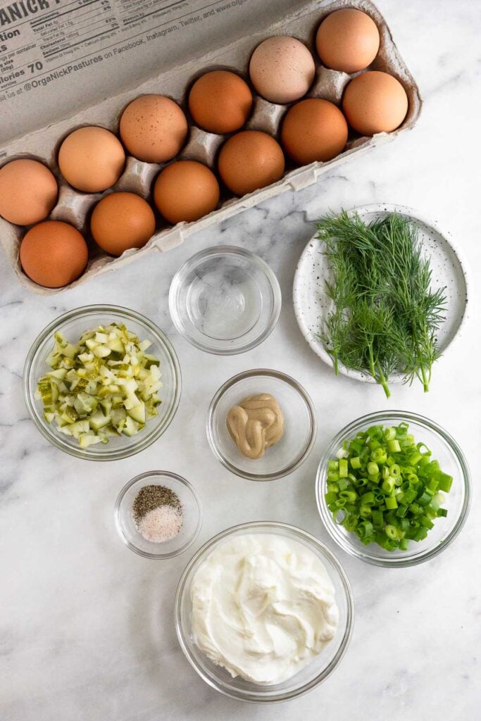 Marble counter with a carton of brown eggs, a plate of fresh dill, a bowl of sliced green onions, a bowl of greek yogurt, a bowl of salt and pepper, a bowl of mustard, a bowl of diced pickles, and a bowl of pickle juice.