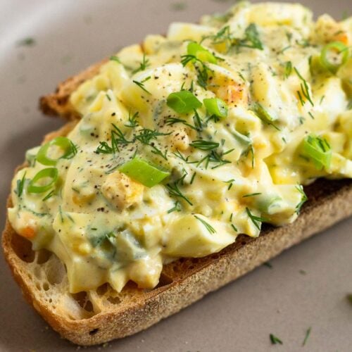 A close up of healthy egg salad on a piece of toast on a brown plate. It is topped with sliced green onions, dill, and black pepper.