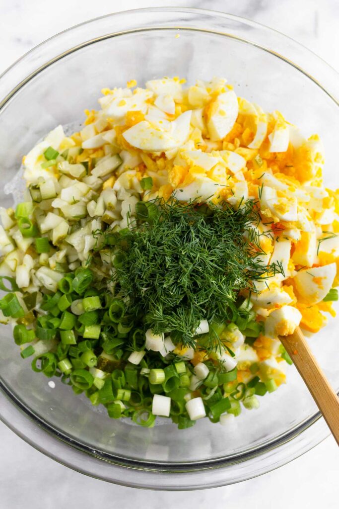Large glass bowl with chopped hard-boiled eggs, diced pickles, sliced green onions, fresh dill, salt, and pepper overtop a greek yogurt mixture. A spatula is in the bowl.