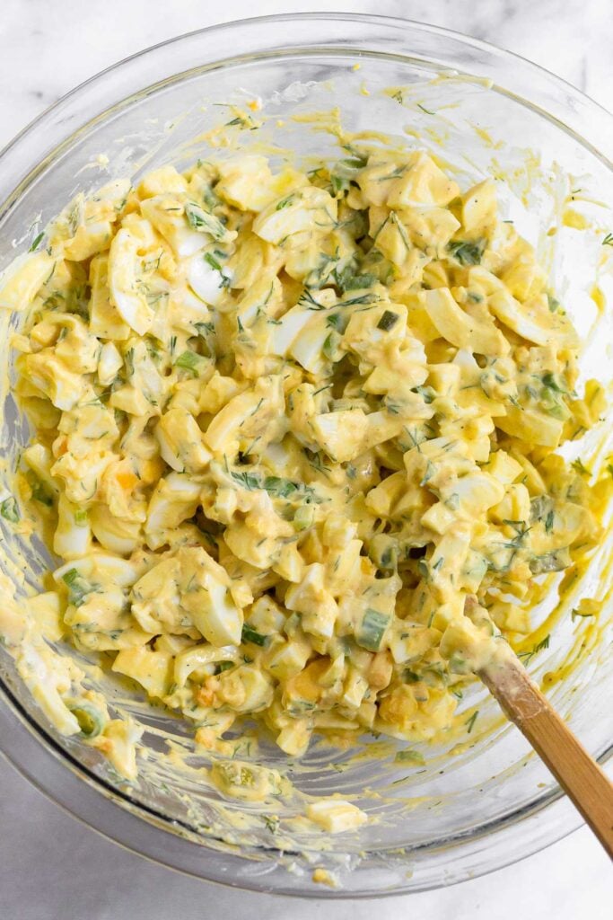 Egg salad with greek yogurt in a large glass bowl with a spatula in it.