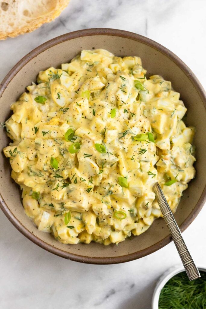 Healthy egg salad in a large brown bowl with a spoon in it. It is topped with dill, green onions, and pepper. Around the bowl is a slice of bread and a bowl of fresh dill.