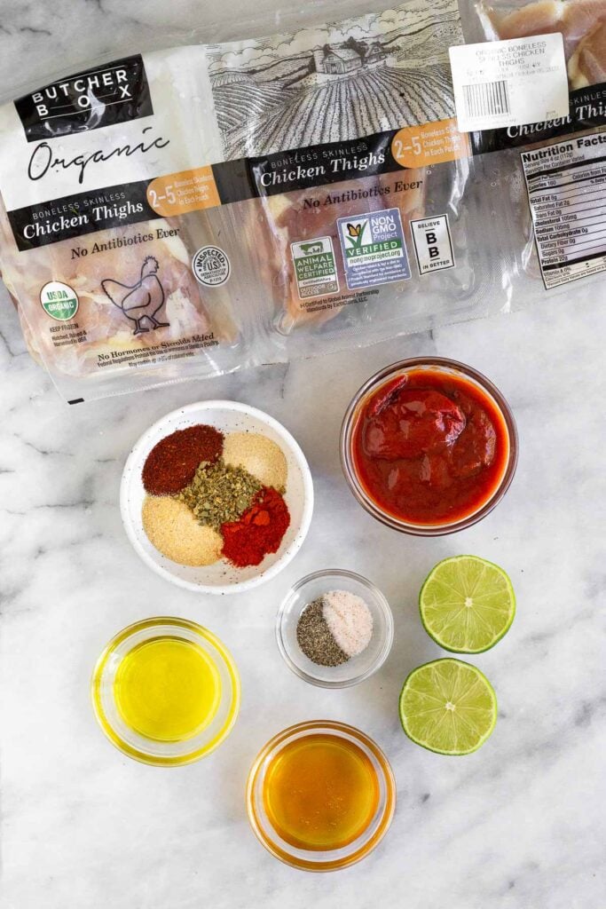 Marble counter with a package of raw chicken thighs, a bowl of chipotle peppers in adobo sauce, a small bowl of salt and pepper, a lime cut in half, a bowl of honey, a bowl of olive oil, and a bowl of spices on it.