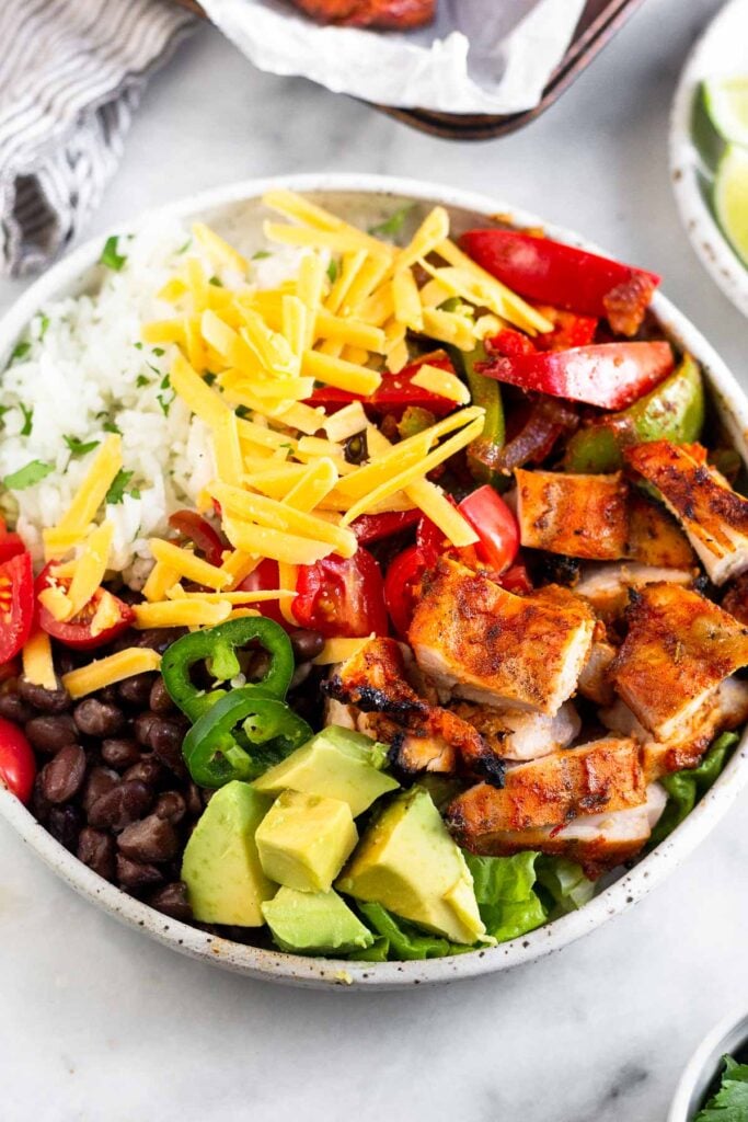 Burrito bowl with cilantro lime rice, fajita veggies, chopped grilled chicken thighs, avocado, sliced jalapeños, black beans, lettuce, tomato, and cheese.