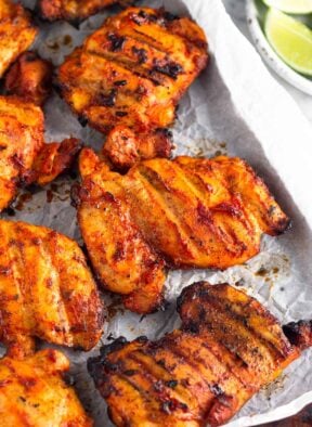 Grilled honey chipotle chicken thighs on a baking sheet lined with parchment paper. Next to it is a plate of lime wedges.