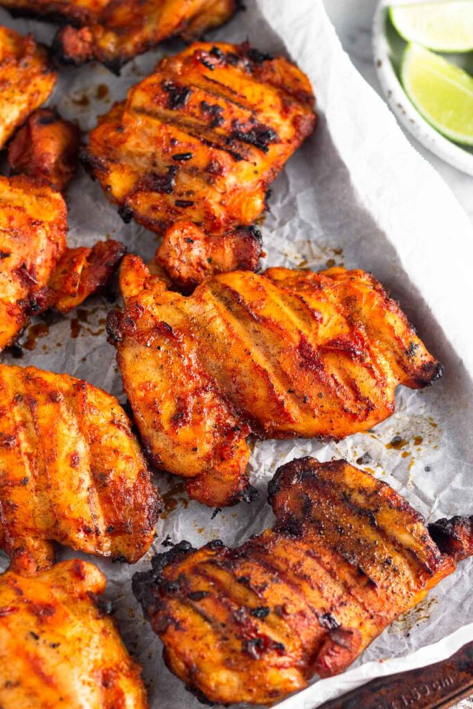Grilled honey chipotle chicken thighs on a baking sheet lined with parchment paper. Next to it is a plate of lime wedges.