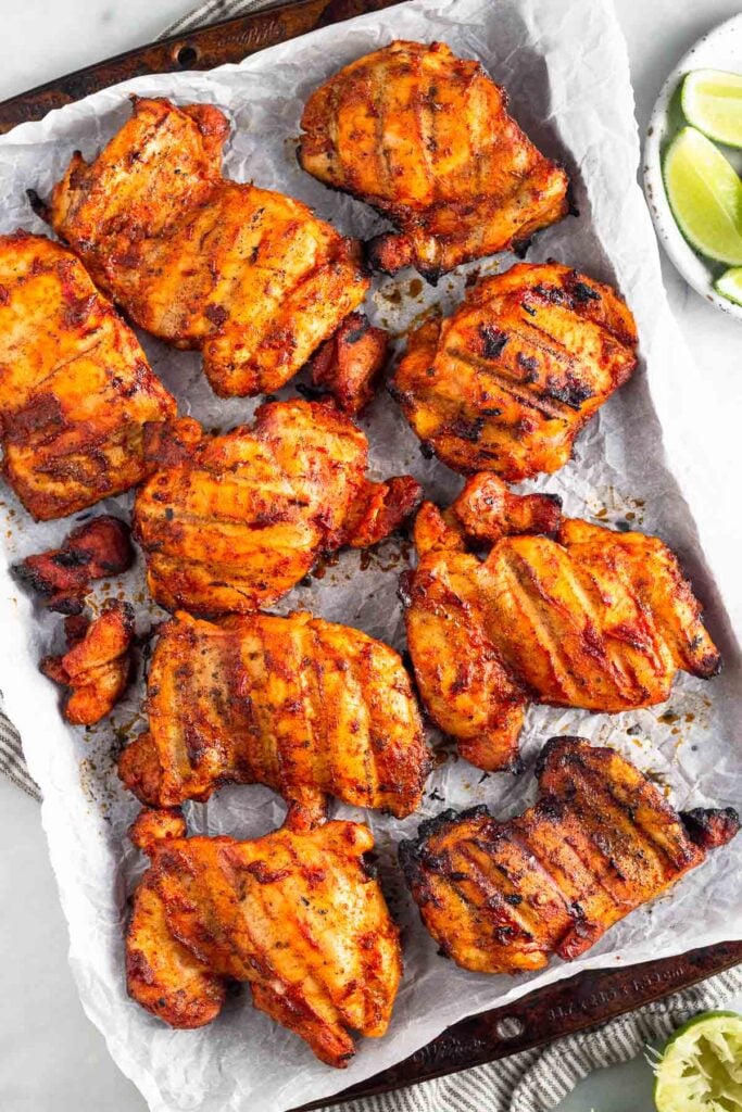 Overhead shot of a baking sheet filled with grilled honey chipotle chicken thighs on it. The baking sheet is on top of a kitchen towel and half a lime and a plate of lime wedges is next to it.