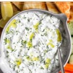 Tzatziki sauce garnished with dill and olive in a large bowl with a spoon in it. It is surrounded by sliced pita bread, sliced cucumber, carrot sticks, and lemon wedges.