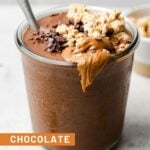 Chocolate protein smoothie in a glass jar with a spoon in it. It is topped with granola, nut butter, and cacao nibs. Around it is some granola and bowl of granola.