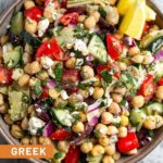Large bowl filled with greek chickpea salad garnished with fresh herbs and two lemon wedges. Next to it is an empty jar and a bowl full of herbs.