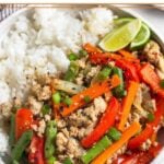 A plate of turkey stir fry with veggies, white rice, and a lime wedge. It is garnished with sesame seeds and green onions.