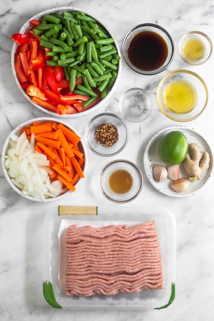 White marble counter top with a bowl of coconut aminos, a bowl of honey, a small bowl of sesame oil, a plate with a lime, 3 cloves of garlic, and fresh ginger on it, a container of raw ground turkey, a bowl of sliced onions and carrots, a small bowl of fish sauce, a small bowl of red pepper flakes, a small bowl of rice vinegar, and a bowl of chopped peppers and green beans.