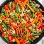 Large cast iron of ground turkey stir fry with peppers, carrots, onions, and green beans. It is topped with green onions and sesame seeds. Next to the skillet is a plate of lime wedges and a bowl of diced green onions.