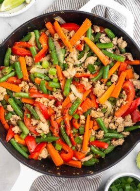 Large cast iron of ground turkey stir fry with peppers, carrots, onions, and green beans. It is topped with green onions and sesame seeds. Next to the skillet is a plate of lime wedges and a bowl of diced green onions.