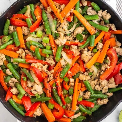 Large cast iron of ground turkey stir fry with peppers, carrots, onions, and green beans. It is topped with green onions and sesame seeds. Next to the skillet is a plate of lime wedges and a bowl of diced green onions.