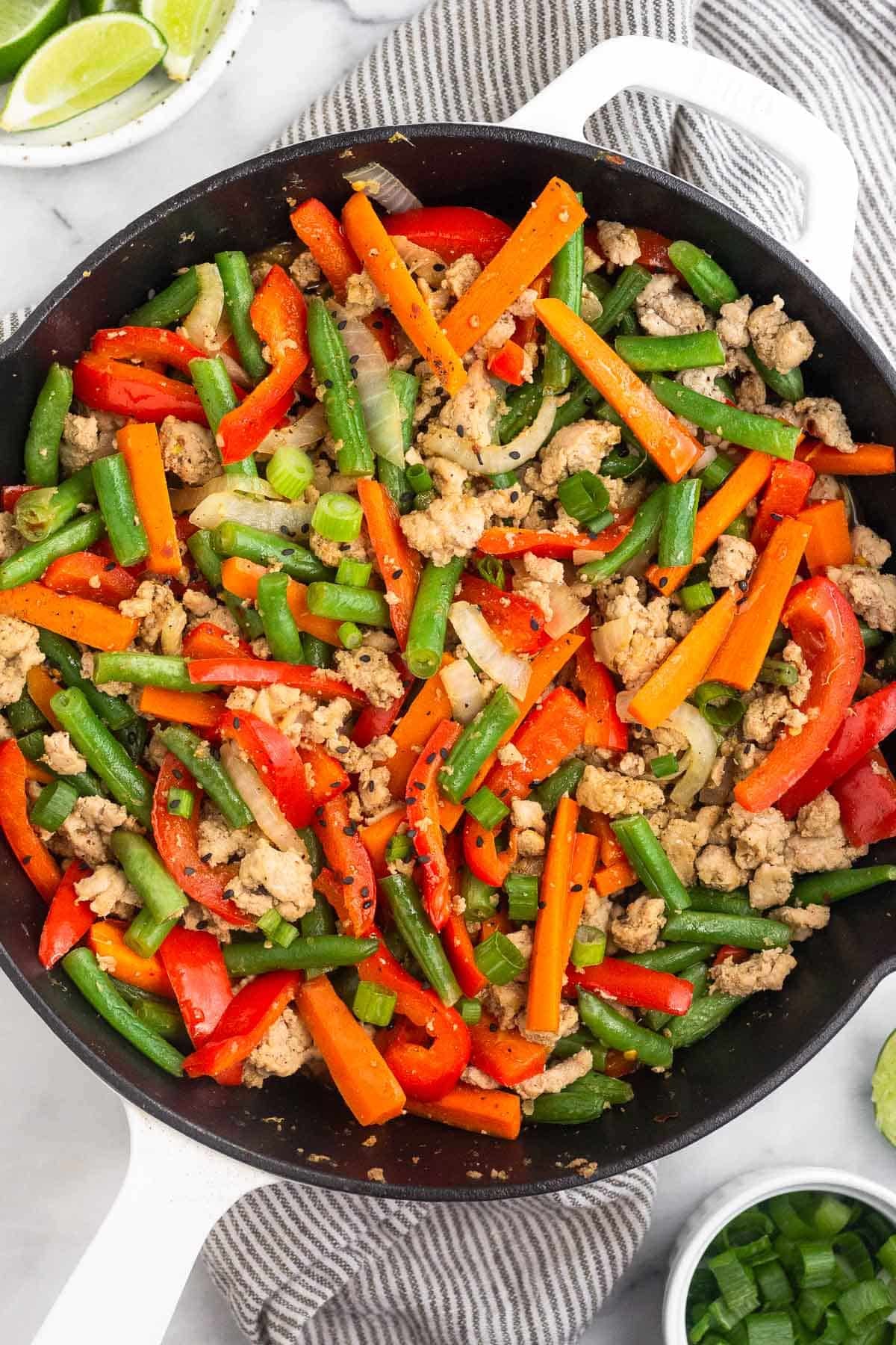 Large cast iron of ground turkey stir fry with peppers, carrots, onions, and green beans. It is topped with green onions and sesame seeds. Next to the skillet is a plate of lime wedges and a bowl of diced green onions.