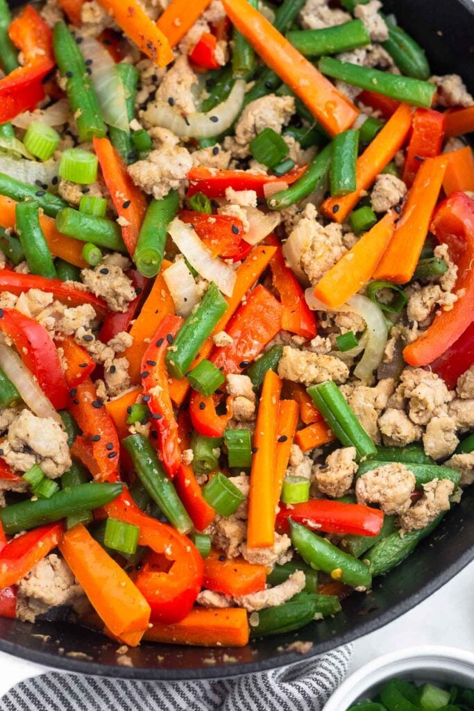 Turkey stir fry with carrots, peppers, onion, and green beans in a large cast iron skillet. It is garnished with green onions and sesame seeds.
