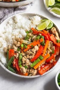 A plate of turkey stir fry with veggies, white rice, and a lime wedge. It is garnished with sesame seeds and green onions. Behind the plate is a plate of lime wedges and a large skillet of more stir fry.