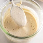 Healthy caesar dressing in a jar with a spoon coming out of it. In front of the jar is half a juiced lemon and behind it is a bowl of salad.