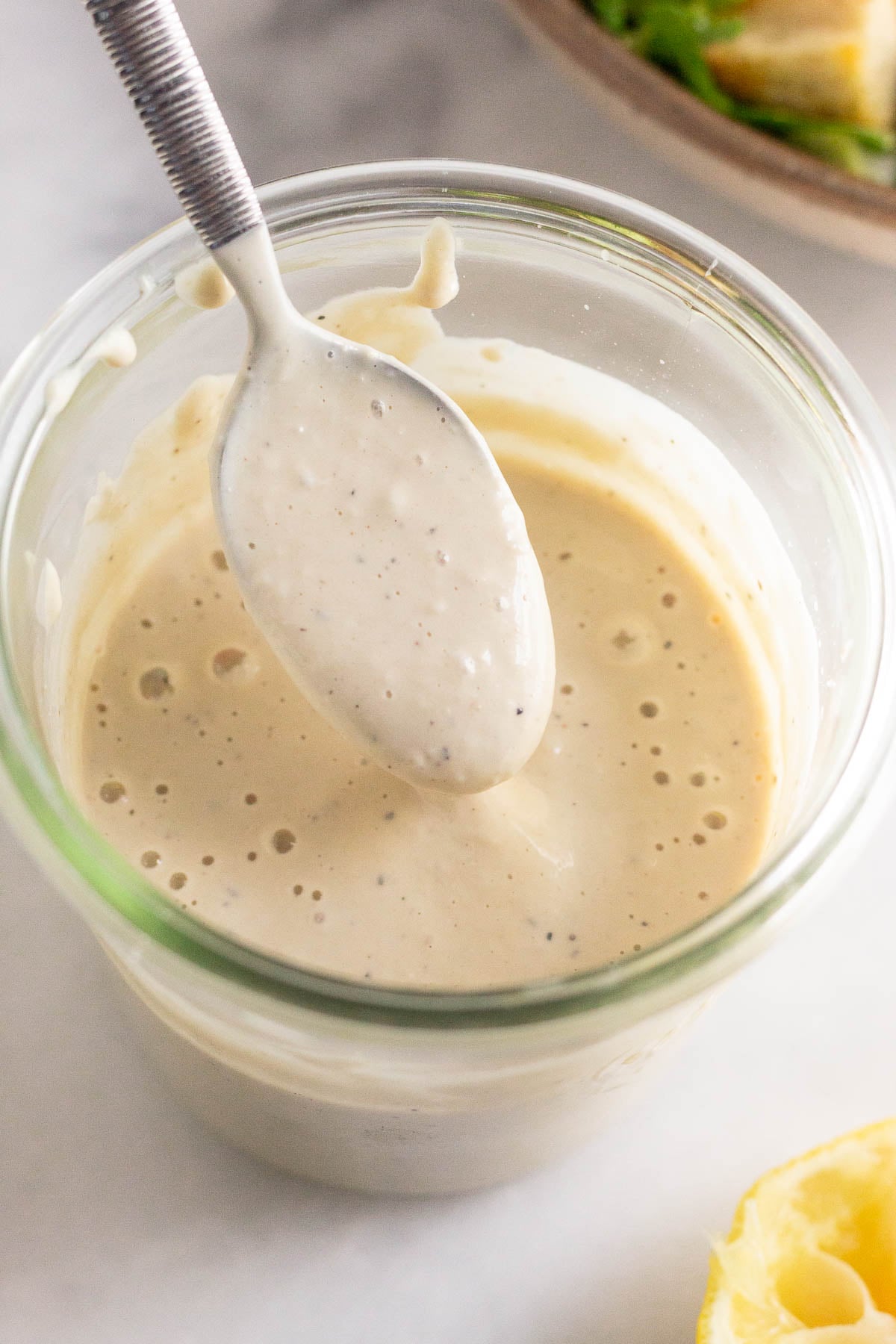 Healthy caesar dressing in a jar with a spoon coming out of it. In front of the jar is half a juiced lemon and behind it is a bowl of salad.