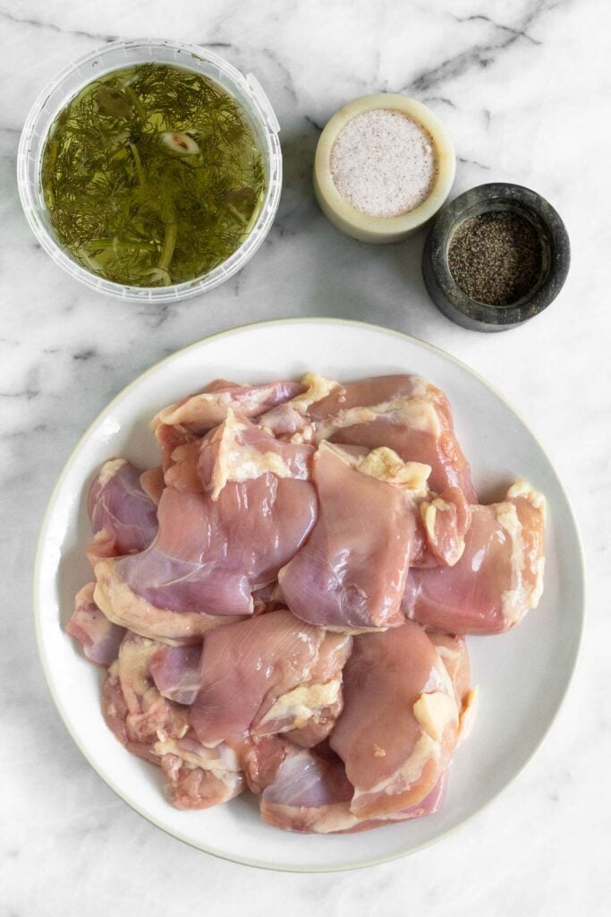 A container of pickle juice, a dish of salt, a dish of pepper, and a plate of raw chicken thighs on a white marble counter.