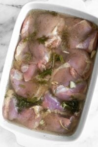A large casserole dish with chicken marinating in pickle juice with garlic and dill.