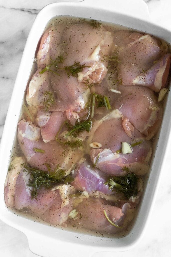 A large casserole dish with chicken marinating in pickle juice with garlic and dill.