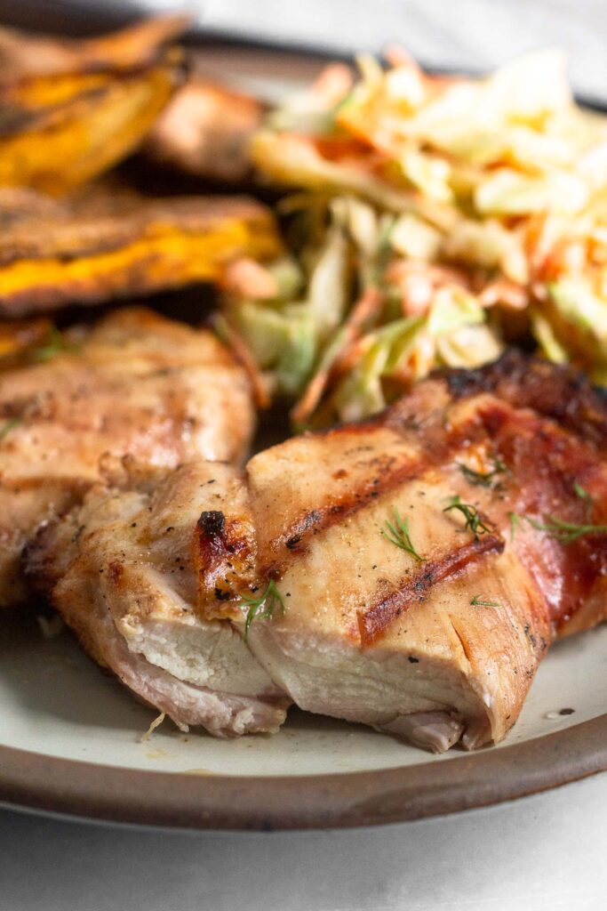 Close up of pickle chicken on a plate with sweet potato fries and coleslaw behind it. A piece is cut off from the chicken.