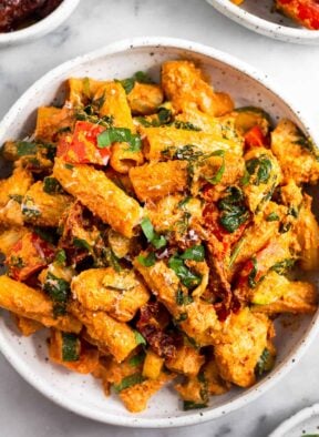 Chicken protein pasta recipe with chicken, veggies, and a sun-dried tomato sauce. Around it is another plate of pasta, a dish of herbs, and a dish of sun-dried tomatoes.