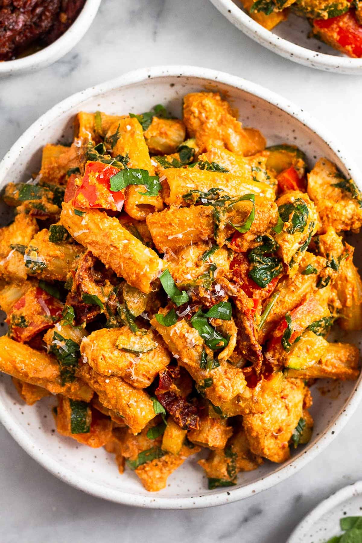 Chicken protein pasta recipe with chicken, veggies, and a sun-dried tomato sauce. Around it is another plate of pasta, a dish of herbs, and a dish of sun-dried tomatoes.