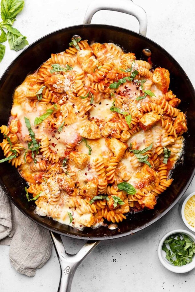 Chicken parmesan pasta in a large skillet topped with red pepper flakes and fresh basil. Next to it is more fresh basil, a kitchen towel, a bowl of chopped basil, and a bowl of parmesan cheese.