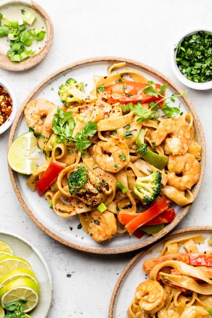 A plate of shrimp and veggies tossed with noddles and sauce. Next to it is another plate, a plate of sliced slimes, and some bowls of herbs and spices.