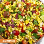 Summer corn salad in a large bowl with fresh corn kernels, diced tomato, diced avocado, diced red onion, and cilantro. The bowl is sitting on a striped towel and around it is some lime wedges, a bunch of cilantro, and some cherry tomatoes.