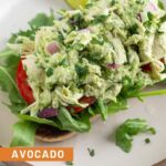 Avocado chicken salad on top of some sliced tomatoes and arugula on a piece of toast on a plate. On the plate is a lime wedge.