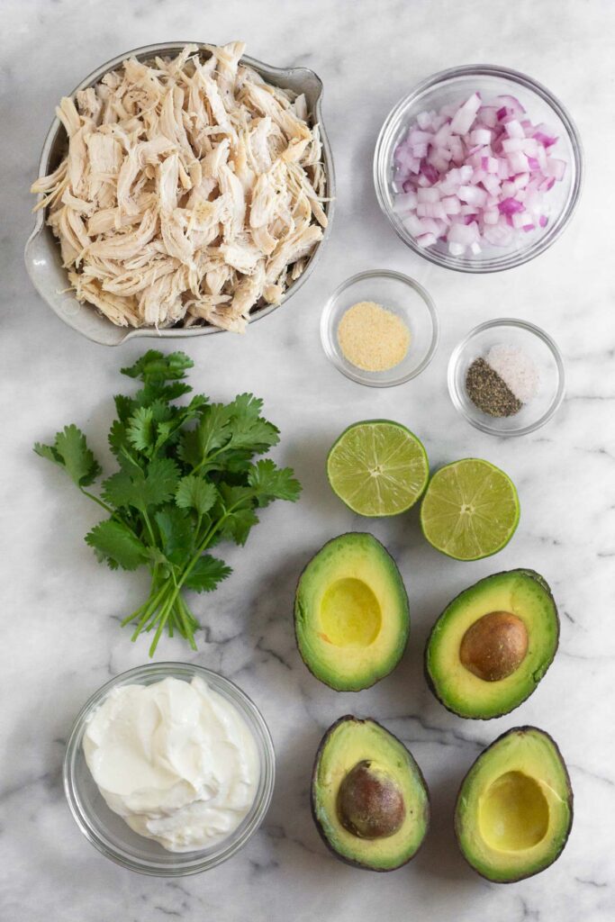 White marble counter with a bowl of shredded chicken, a bowl of diced red onions, two small bowls of spices, a lime cut in half, 4 halves of avocados, a bowl of yogurt, and a bunch of cilantro.