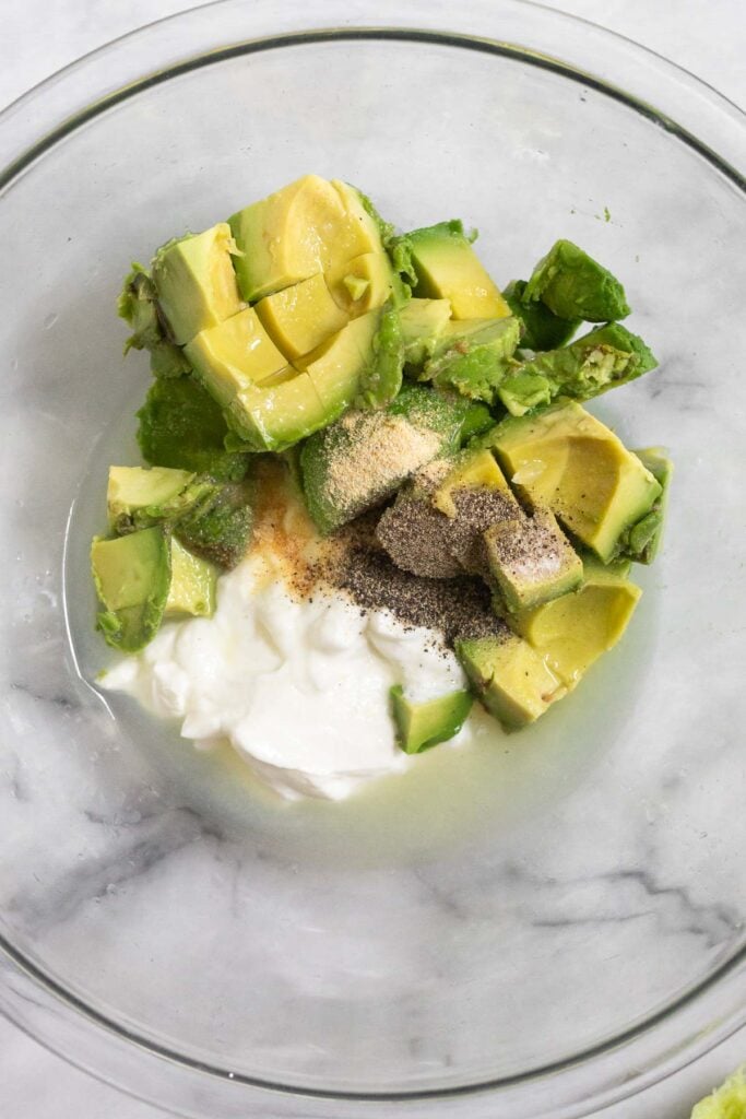 Glass bowl with greek yogurt, diced avocados, lime juice, and spices.