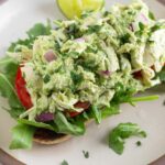 Avocado chicken salad on top of some sliced tomatoes and arugula on a piece of toast on a plate. On the plate is a lime wedge.