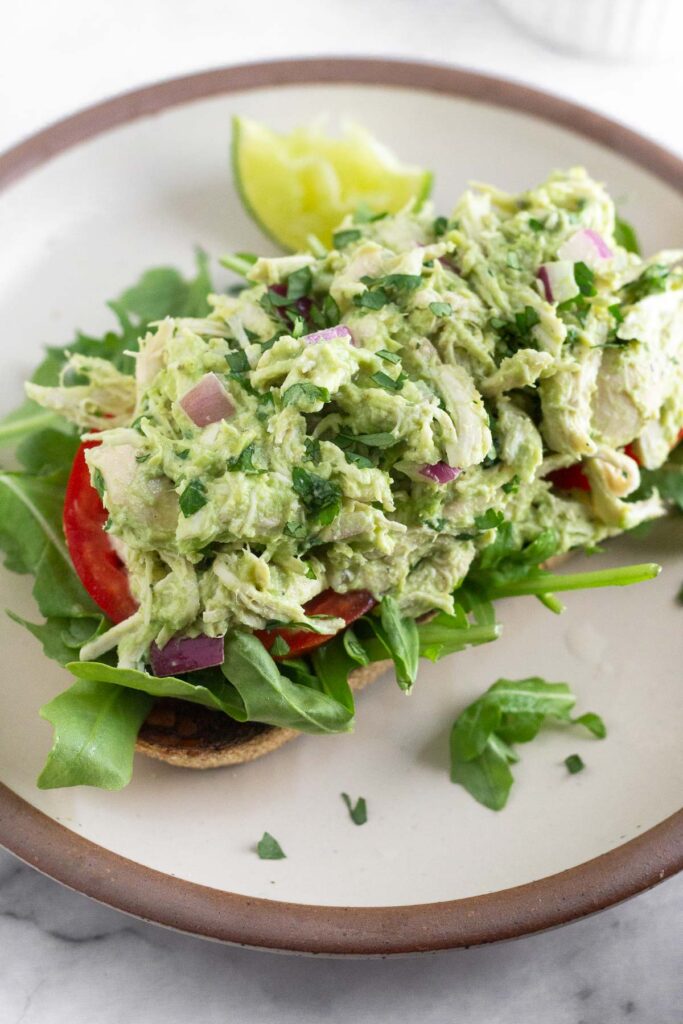Avocado chicken salad on top of some sliced tomatoes and arugula on a piece of toast on a plate. On the plate is a lime wedge.