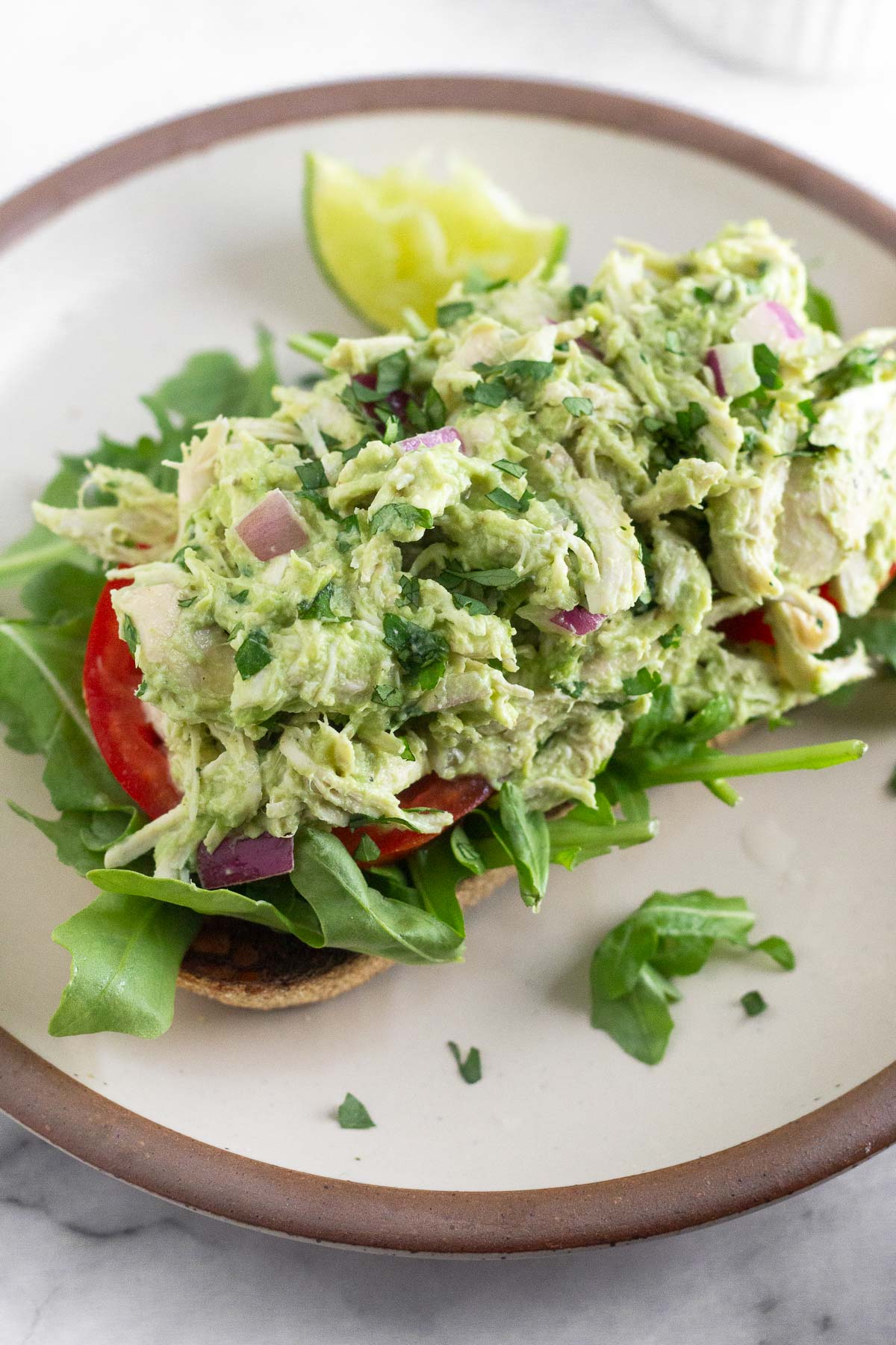 Avocado chicken salad on top of some sliced tomatoes and arugula on a piece of toast on a plate. On the plate is a lime wedge.