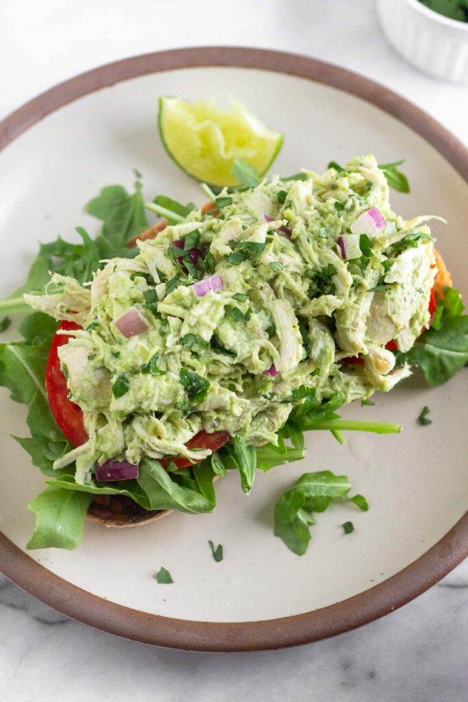 Overhead shot of avocado chicken salad with on top of some sliced tomatoes and arugula on a piece of toast on a plate. A lime wedge is also on the plate.