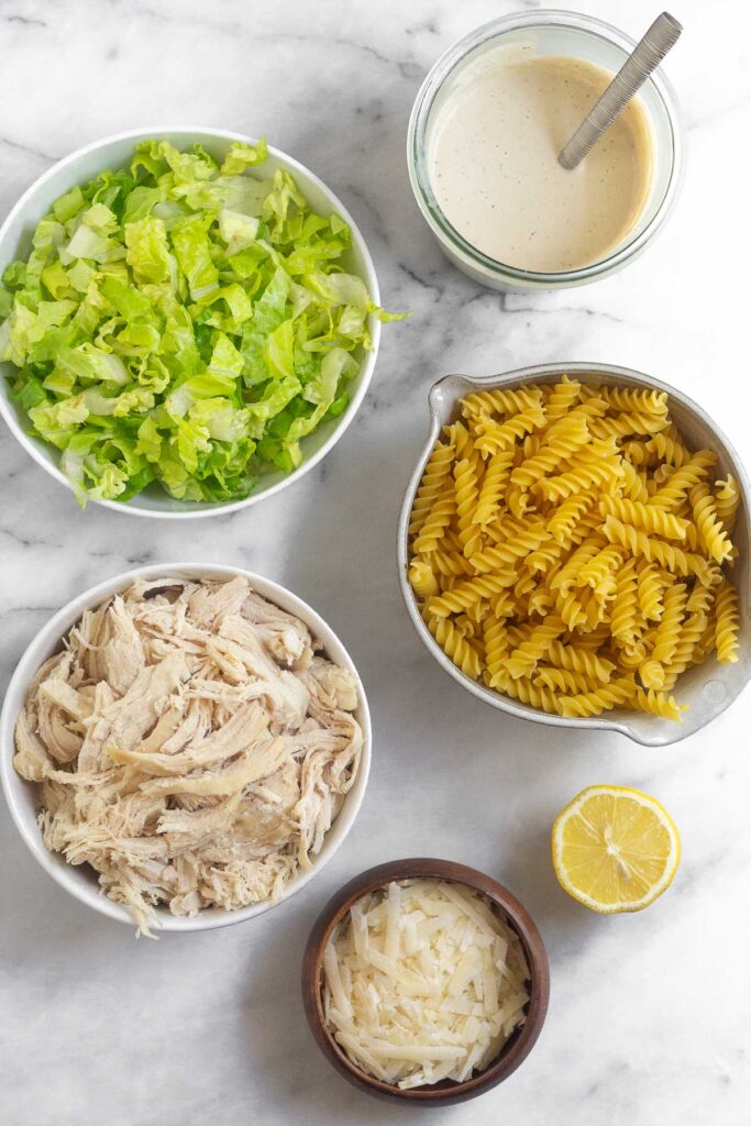 White marble counter with a jar of caesar dressing with a spoon in it, a bowl of uncooked pasta, half a lemon, a bowl of grated parmesan cheese, a bowl of shredded chicken, and a bowl of chopped romaine lettuce.