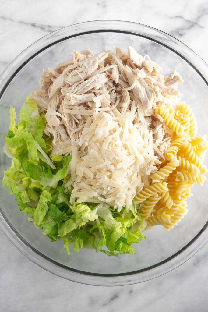 A large glass bowl of cooked pasta, chopped romaine, shredded chicken, and grated parmesan cheese.