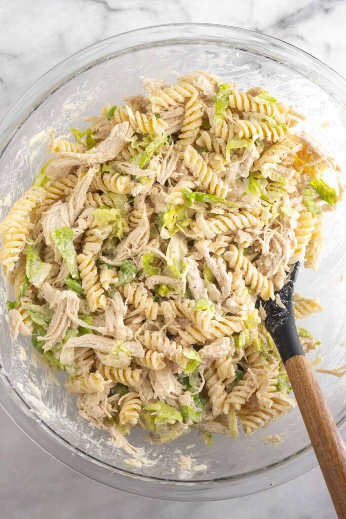 Large glass bowl with shredded chicken, chopped romaine, parmesan cheese, and pasta tossed in a creamy dressing. There is a spoon in the bowl as well.