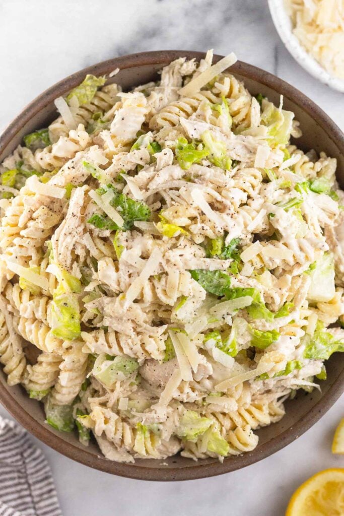 Overhead shot of caesar pasta salad with chicken in a brown bowl. Around the bowl is half a juiced lemon, a striped kitchen bowl, and a bowl of shredded parmesan cheese.
