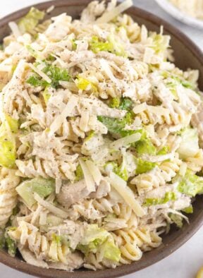 A close up of a bowl of chicken caesar pasta salad. It is garnished with black pepper. Behind the bowl is a plate of grated parmesan cheese.