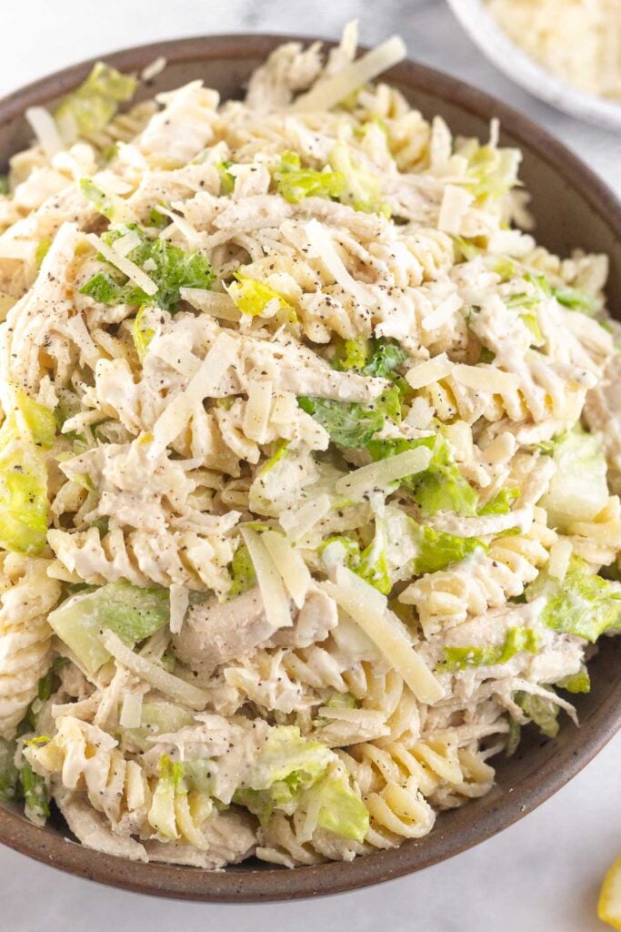 A close up of a bowl of chicken caesar pasta salad. It is garnished with black pepper. Behind the bowl is a plate of grated parmesan cheese.
