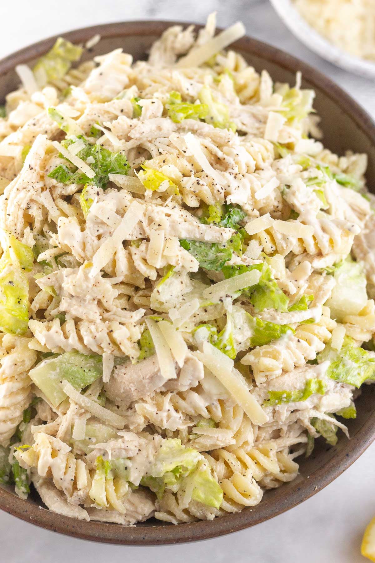 A close up of a bowl of chicken caesar pasta salad. It is garnished with black pepper. Behind the bowl is a plate of grated parmesan cheese.