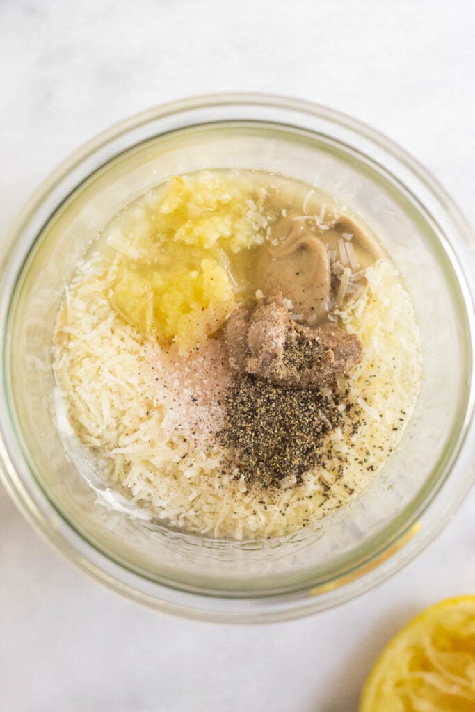 Overhead shot of a large jar with tahini, grated parmesan cheese, minced garlic, dijon mustard, anchovy paste, salt, and pepper in it. Next to is it a half a juiced lemon.